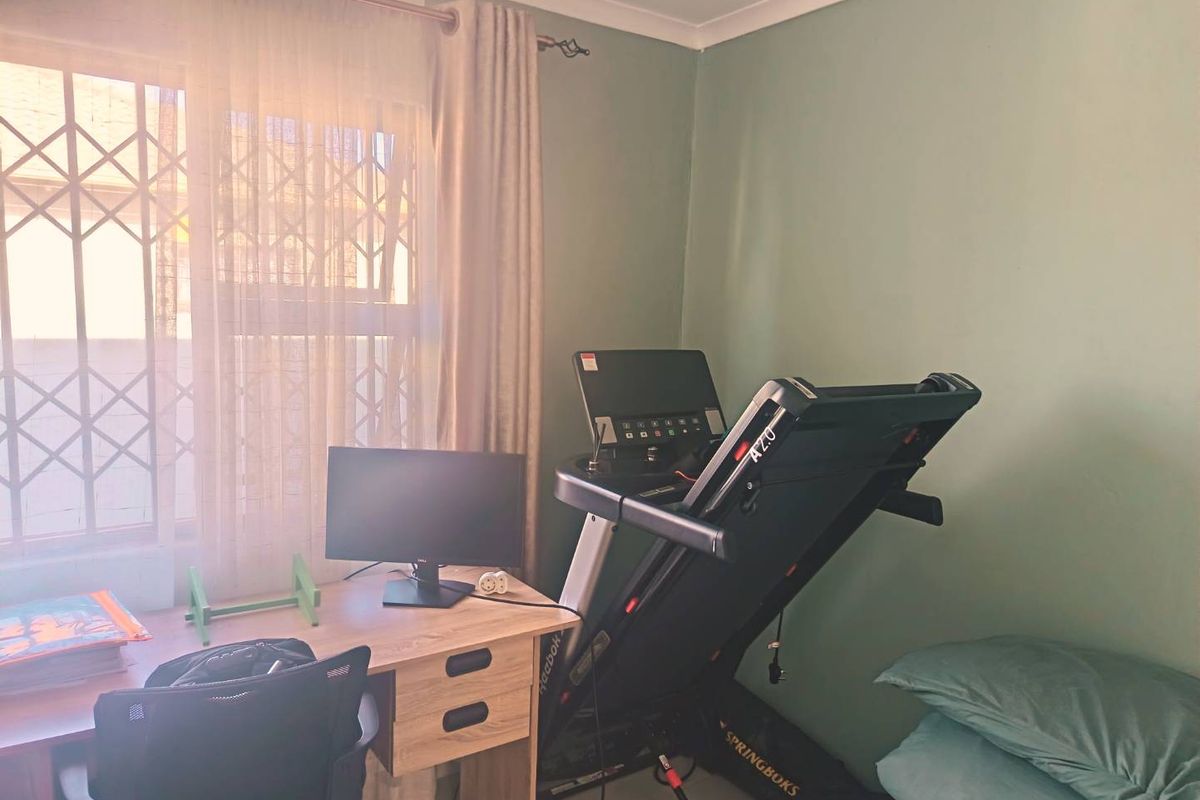 A bedroom currently utilized as a home office and gym space, featuring a window with a security gate and neutral wall finishes.