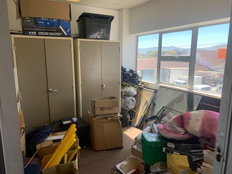 A cluttered office space featuring metal storage cabinets, large windows providing natural light, and a suspended ceiling.