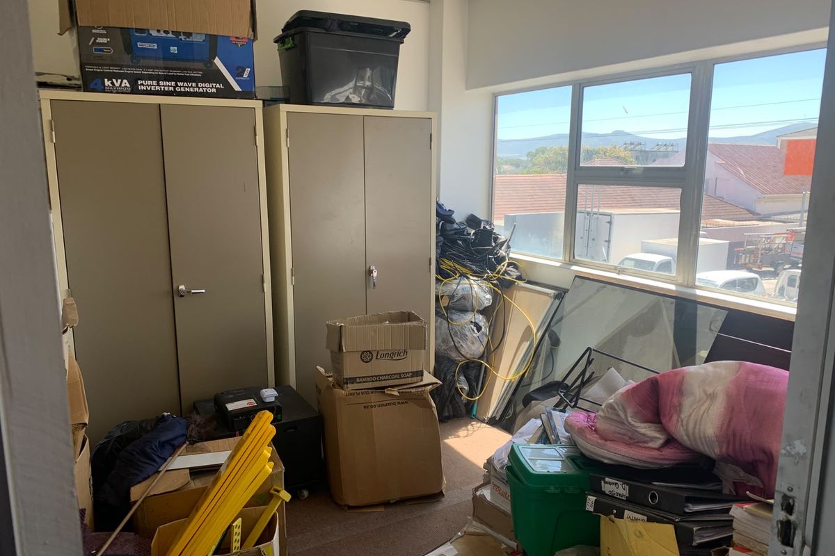 A cluttered office space featuring metal storage cabinets, large windows providing natural light, and a suspended ceiling.