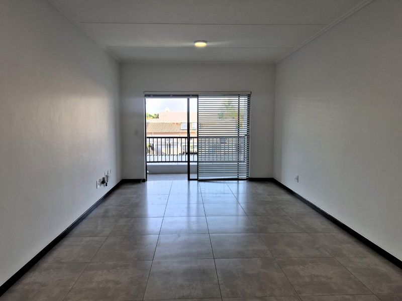 A bright, empty living room featuring neutral tiled flooring, clean white walls, and a sliding glass door leading to a balcony.