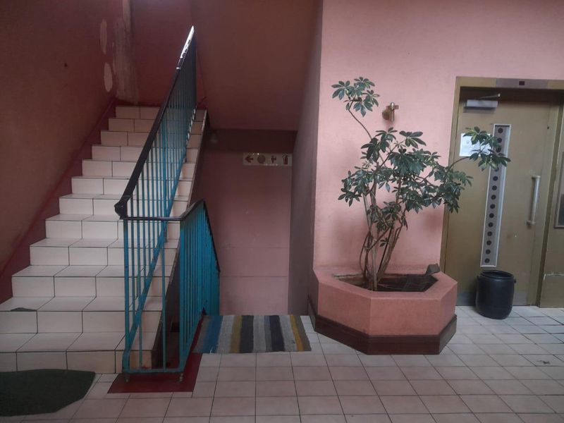 A communal apartment hallway featuring a tiled staircase with metal balustrade, an elevator, and a built-in planter, showing signs of wear and dated finishes.