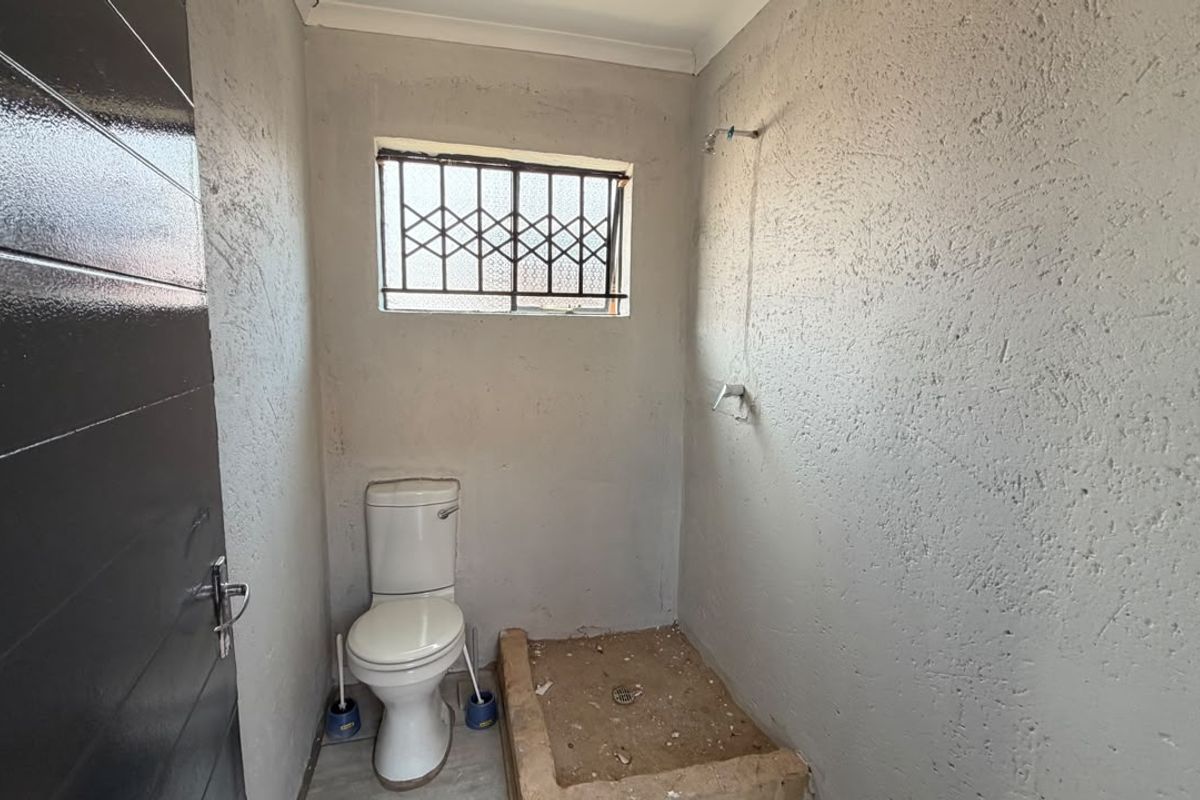 A compact, unfinished bathroom featuring a toilet and a basic shower area with a raised base and security-barred window.