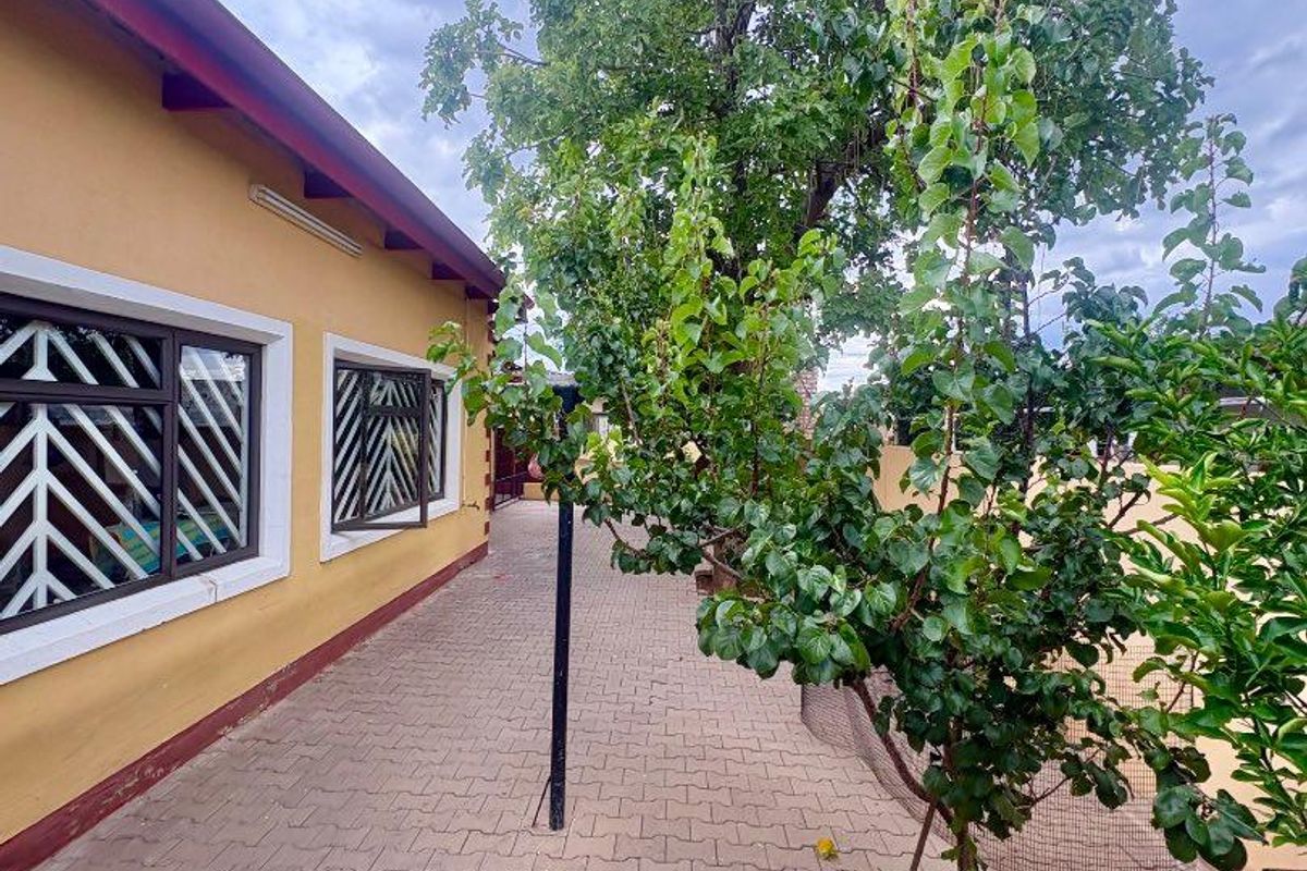 A paved side walkway along the exterior of the house, featuring security bars on windows and mature trees.