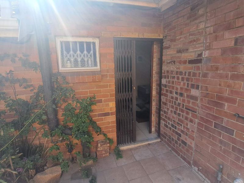 A rear exterior view showing a brick facade, a security gate entrance, and a small tiled patio area.