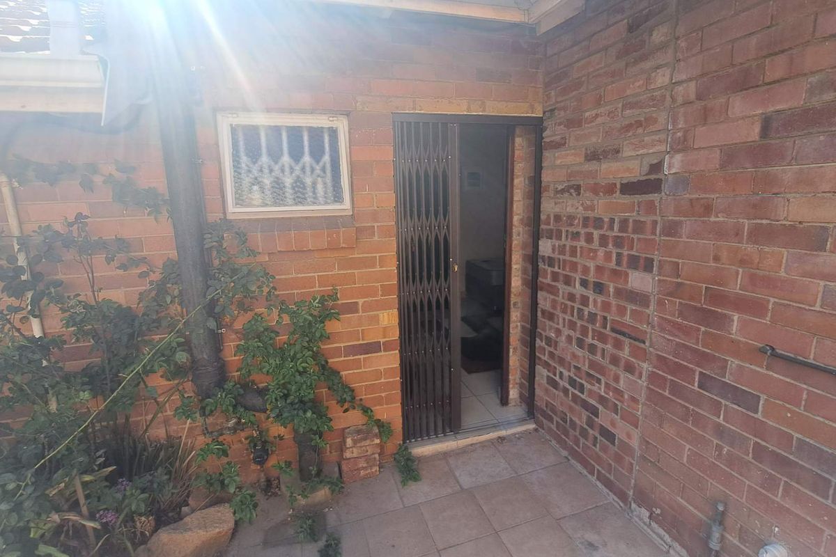 A rear exterior view showing a brick facade, a security gate entrance, and a small tiled patio area.