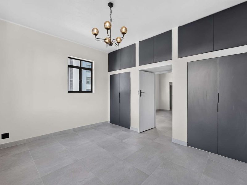 A bright, modern bedroom featuring neutral tiled flooring, built-in dark grey cabinetry, and a contemporary chandelier light fixture.