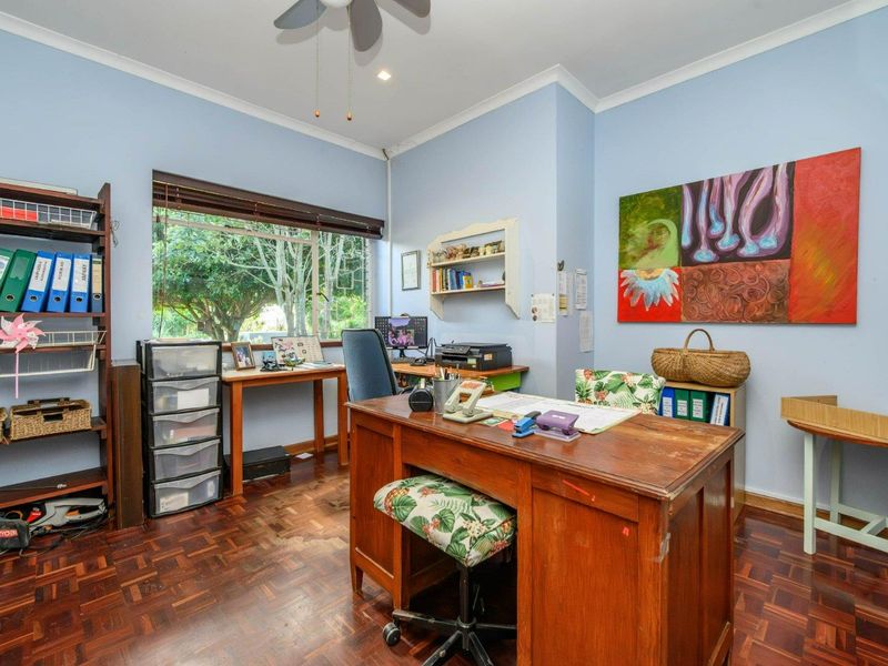 A functional home office featuring classic parquet flooring, a ceiling fan, and large windows providing natural light and garden views.