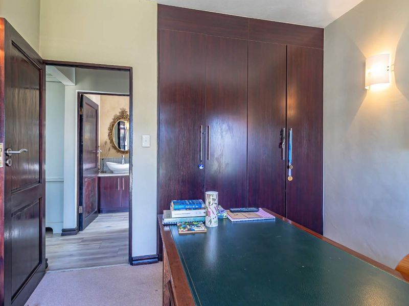 A functional home office featuring dark wood built-in cupboards and wall-mounted lighting, with a view into an adjacent bathroom.