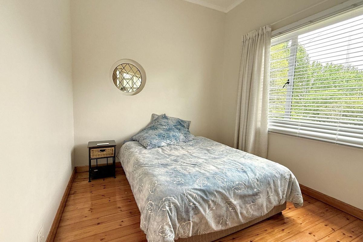 A bright bedroom featuring polished timber flooring, a unique circular accent window, and large window with fitted blinds.