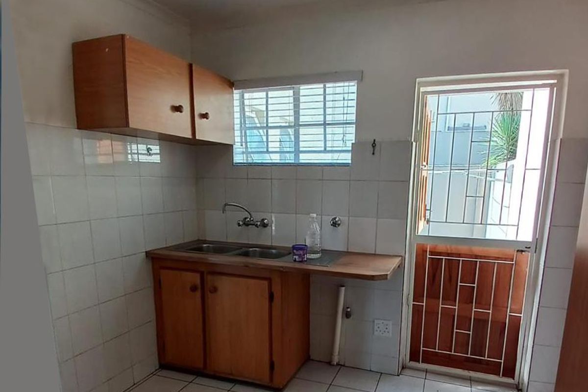A compact kitchen area featuring wooden cabinetry, a double sink, tiled flooring, and direct access to an exterior door with security gate.