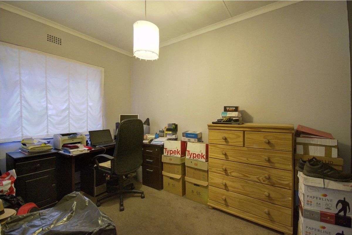 A functional bedroom or home office space featuring neutral wall finishes, carpeted flooring, and a window with a fitted blind.