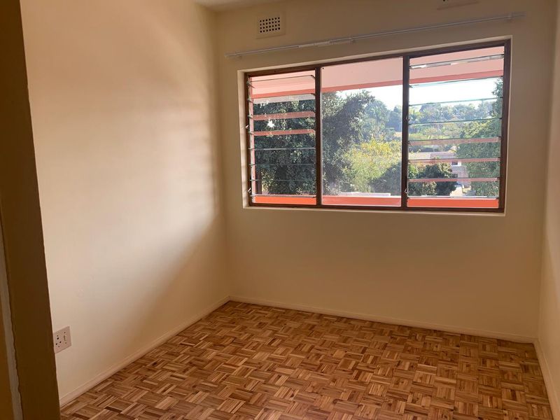 An empty bedroom featuring original parquet flooring and louver-style windows with natural light and a view of the surrounding neighborhood.