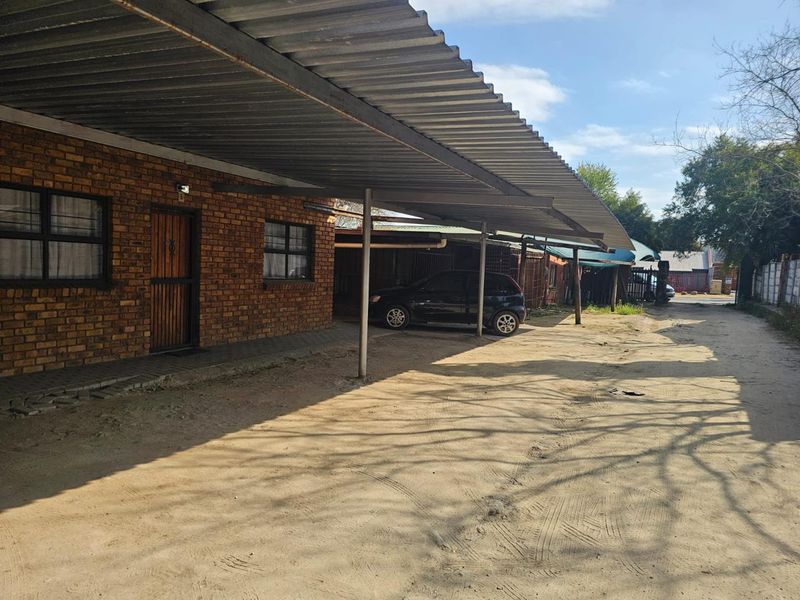 Exterior view of a facebrick residential building featuring a large corrugated metal carport and an unpaved sandy driveway area.