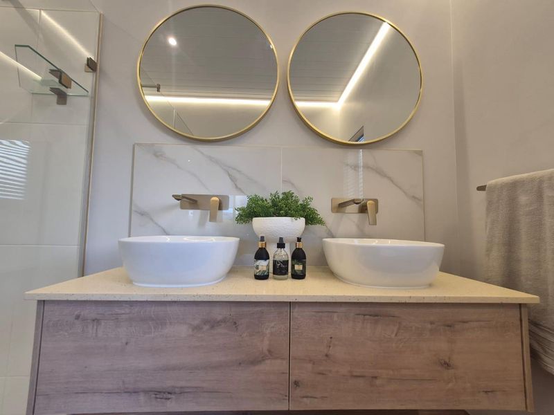 Modern bathroom featuring a double vanity with vessel sinks, wall-mounted gold-tone faucets, marble-look splashback, and round mirrors.