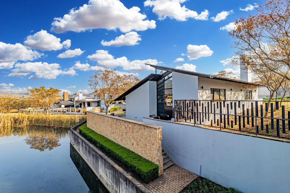 A modern architectural residence featuring a stone retaining wall, metal railings, and a scenic waterfront position overlooking a tranquil pond.