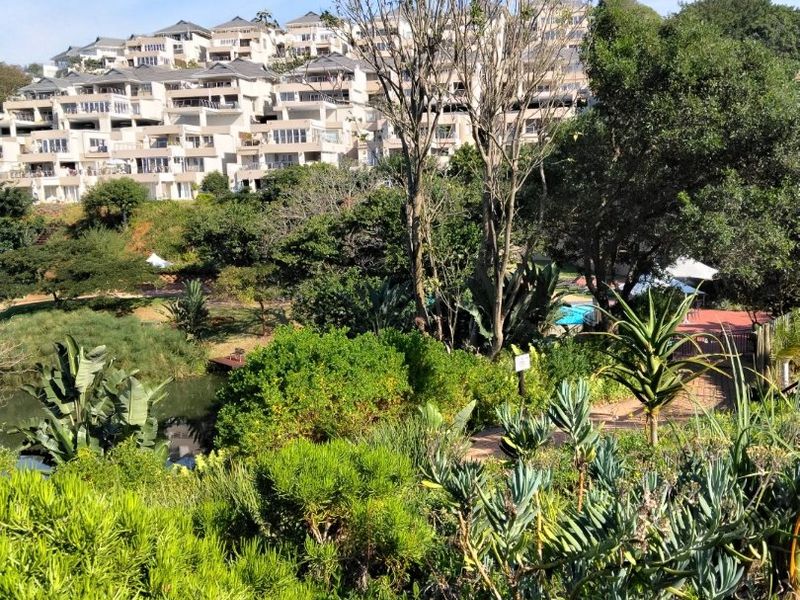 A scenic view of a multi-level apartment complex overlooking a lush, well-maintained garden area with mature trees and native landscaping.