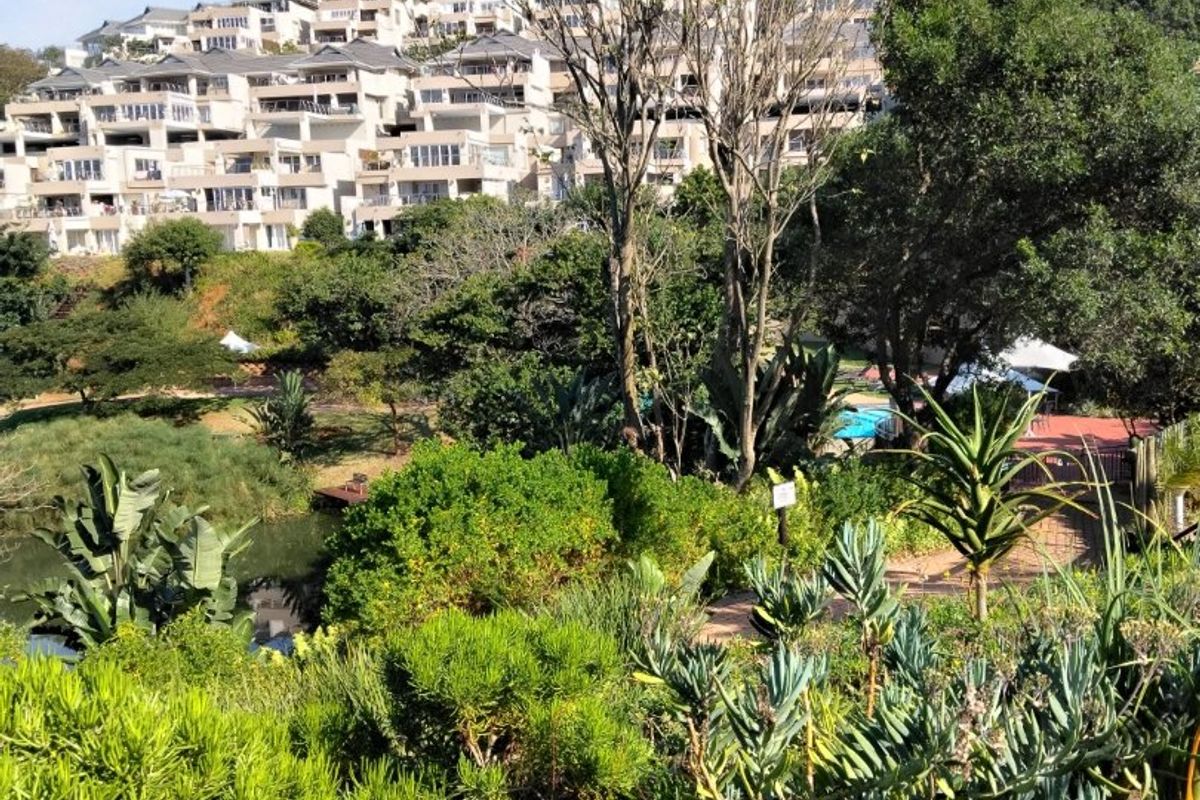 A scenic view of a multi-level apartment complex overlooking a lush, well-maintained garden area with mature trees and native landscaping.