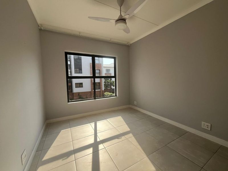 A bright, empty bedroom featuring neutral grey walls, large aluminium-framed windows, and clean tiled flooring with an installed ceiling fan.