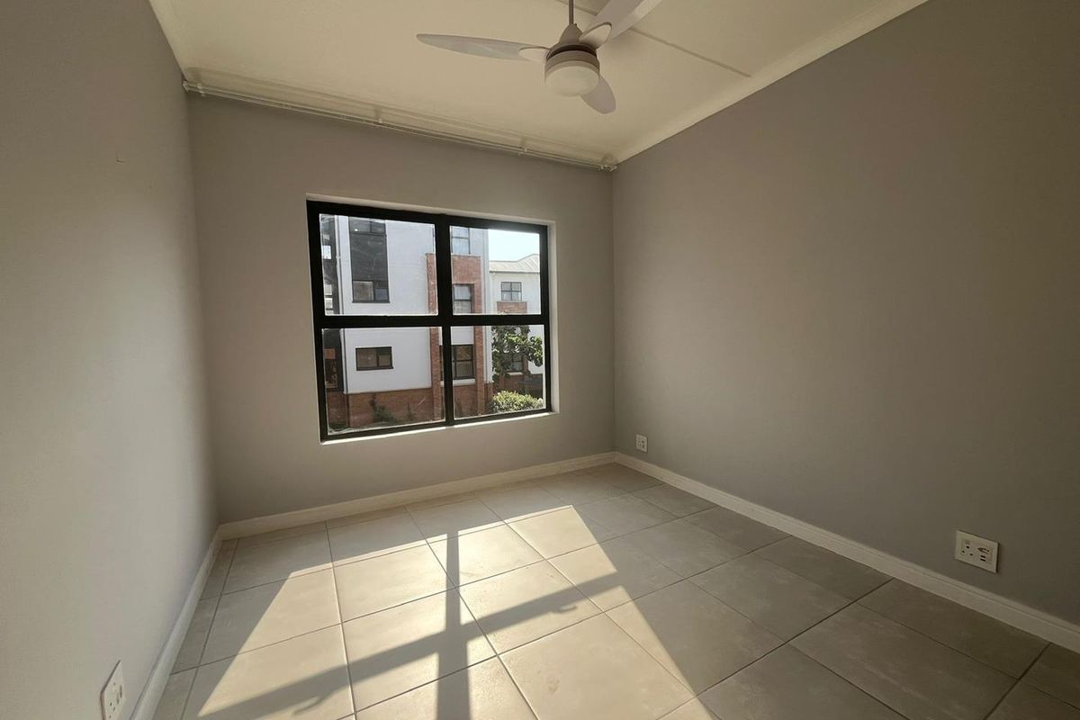 A bright, empty bedroom featuring neutral grey walls, large aluminium-framed windows, and clean tiled flooring with an installed ceiling fan.