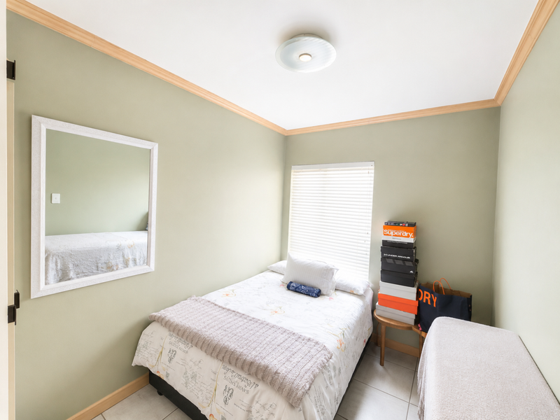 A compact bedroom featuring tiled flooring, neutral green walls with timber-look cornice detailing, and a window fitted with venetian blinds.