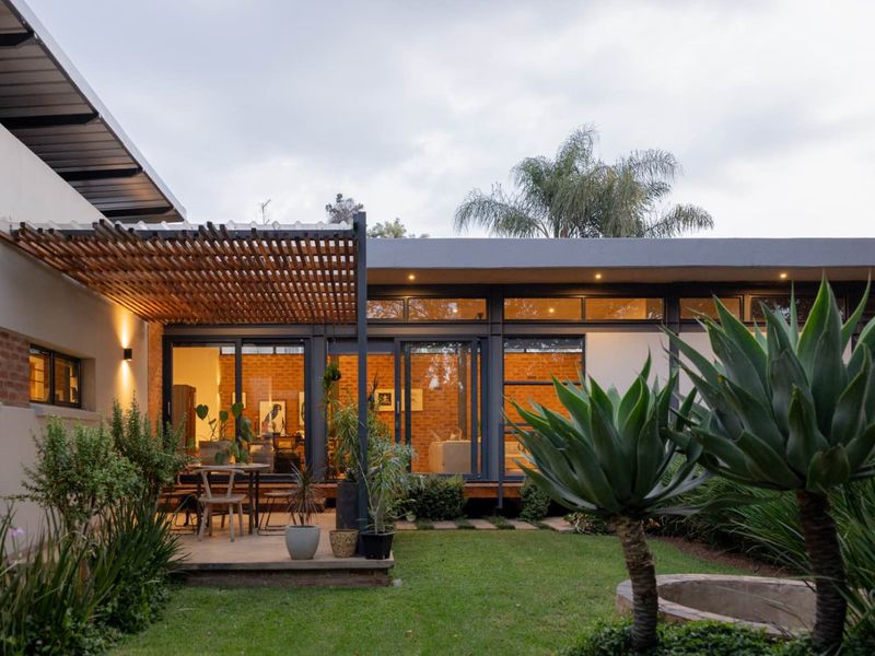A modern rear exterior featuring a timber pergola, concrete patio, and large sliding glass doors opening into a contemporary living space with exposed brick walls.