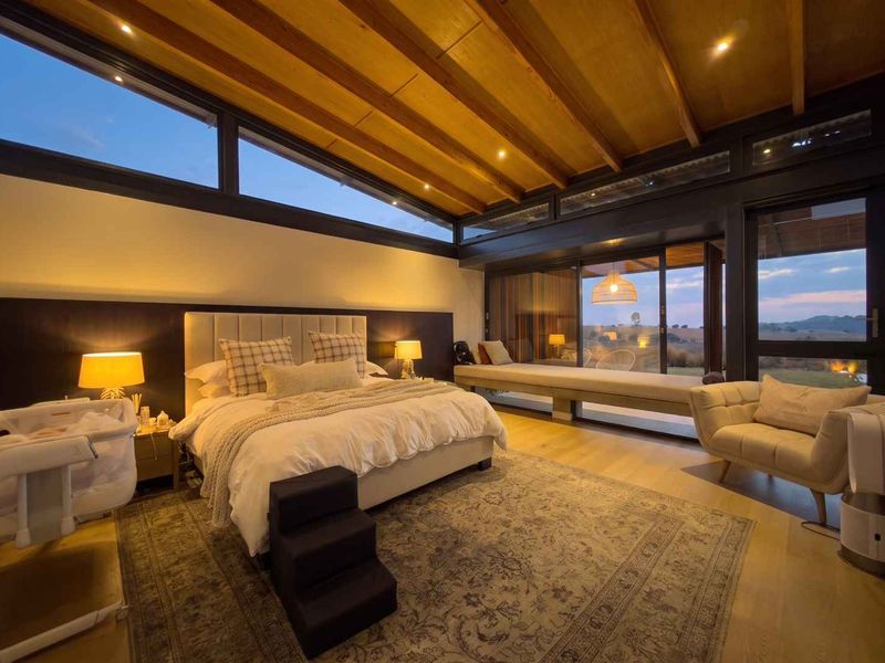 Spacious modern bedroom featuring exposed timber beam ceilings, clerestory windows, and large sliding glass doors that open to scenic views with a built-in window bench.