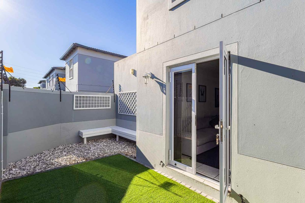 A compact rear courtyard featuring artificial turf, a gravel border, a built-in bench, and a glass door providing access to the interior living space.