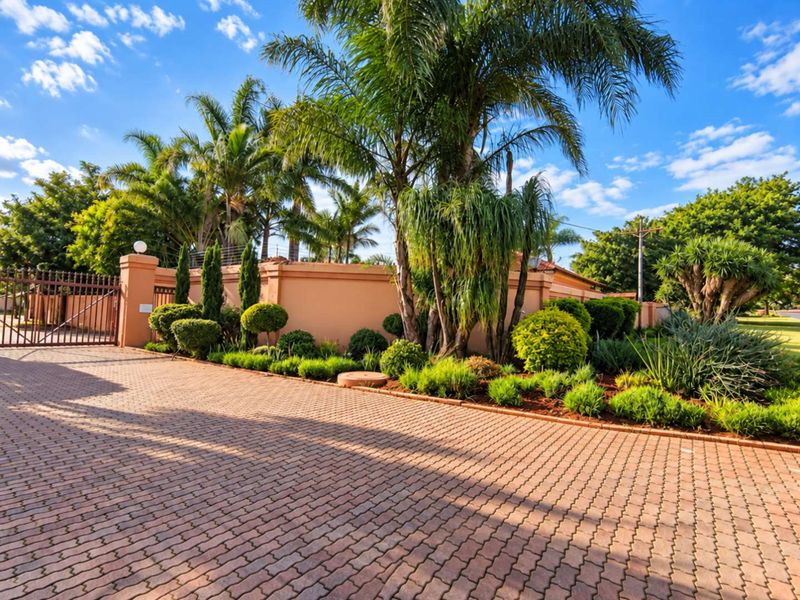 A well-maintained front entrance featuring a paved driveway, secure perimeter wall with metal gate, and established garden landscaping.