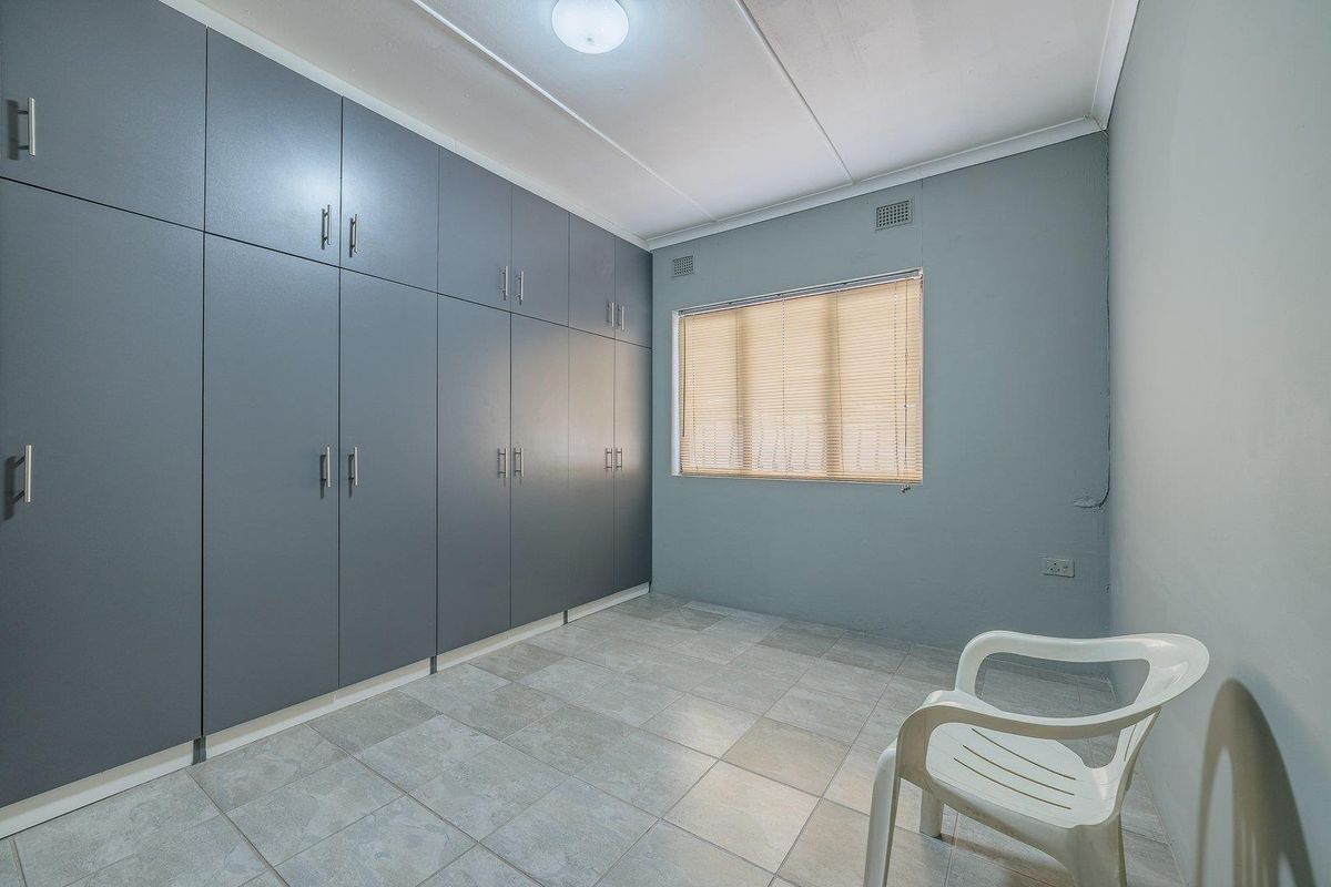 A clean, empty bedroom featuring extensive grey built-in cupboards, tiled flooring, and a window with fitted blinds.