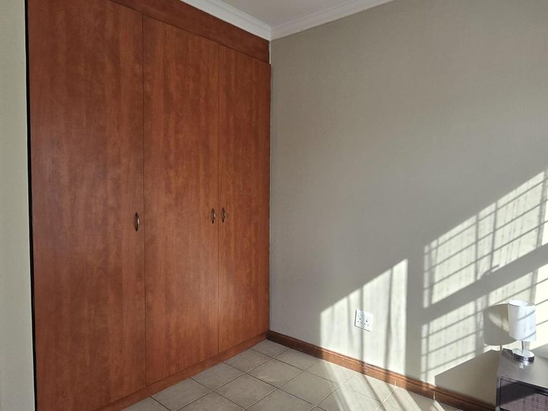 A bright, empty bedroom featuring built-in wooden cupboards, tiled flooring, and natural light streaming through the window.