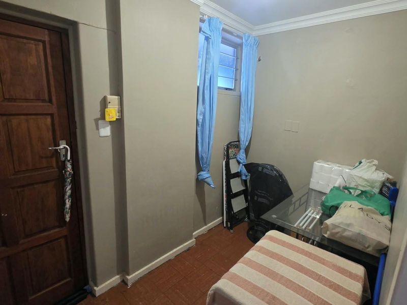 A compact bedroom featuring tiled flooring, a wooden door, and a louvered window, offering a functional space ready for modernization and aesthetic updates.