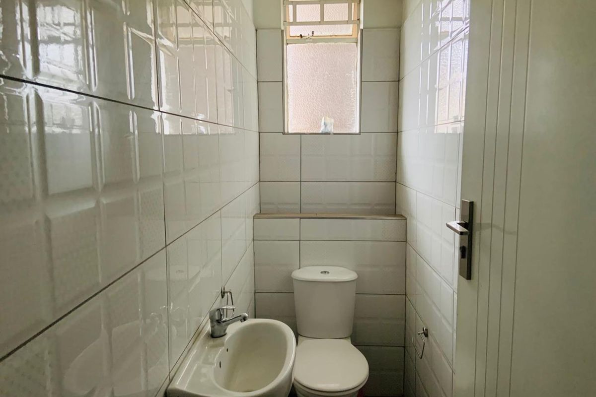 A compact bathroom featuring white wall tiles, a standard toilet, and a small wall-mounted basin with a window for natural light.