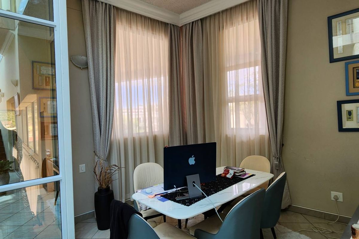 A bright and well-maintained home office featuring large windows, tiled flooring, and a modern glass partition wall overlooking the adjacent hallway.