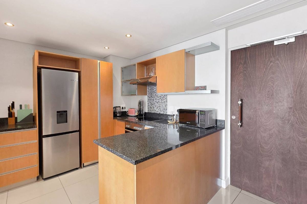 A compact, modern kitchen featuring light wood cabinetry, granite countertops, a mosaic backsplash, and integrated appliances, finished with neutral floor tiles.