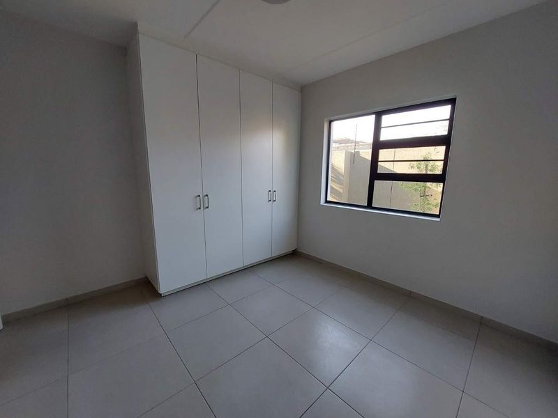 A clean, empty bedroom featuring neutral tiled flooring, built-in white cupboards, and a modern aluminium-framed window.