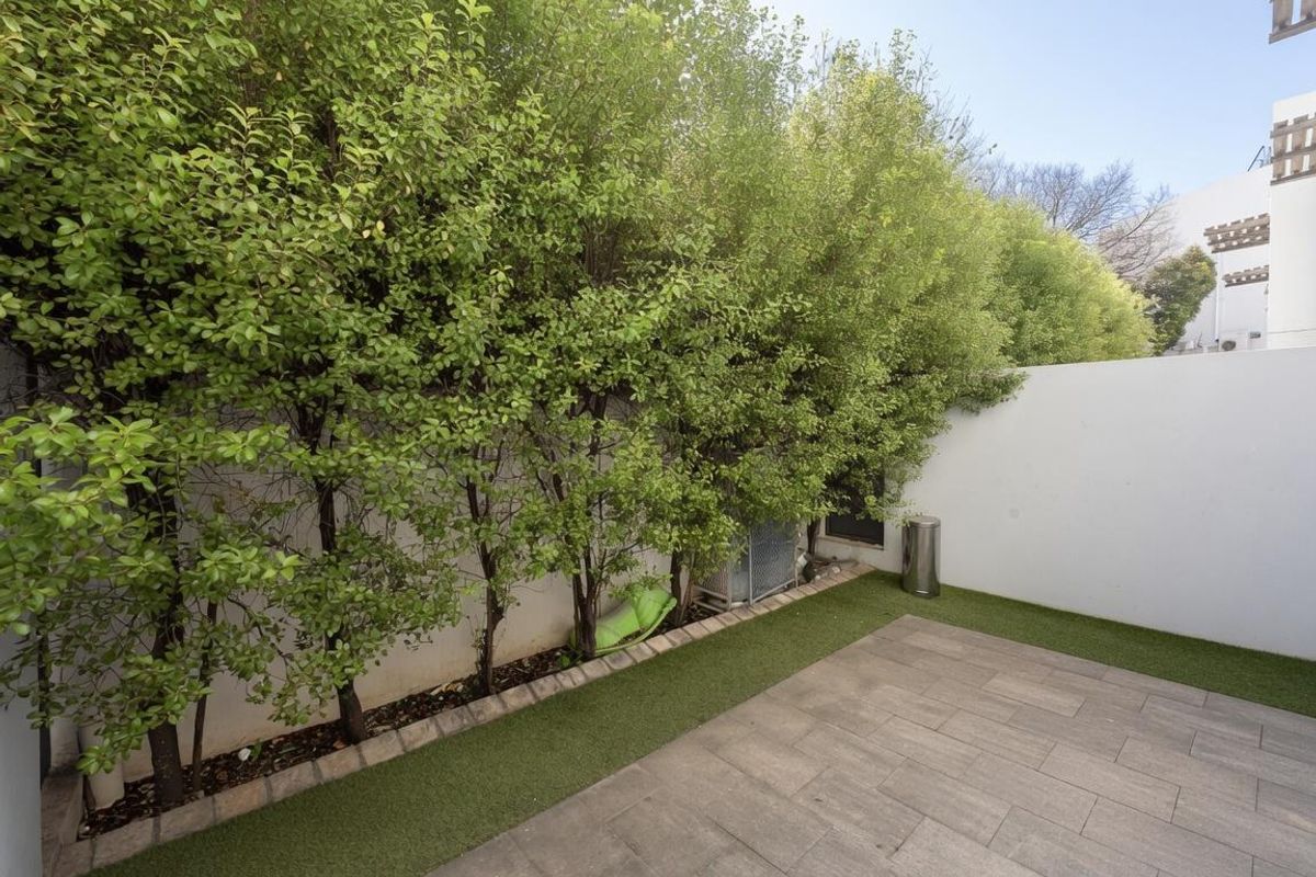 A private rear courtyard featuring paved flooring, artificial grass borders, and established perimeter hedging against a clean white boundary wall.