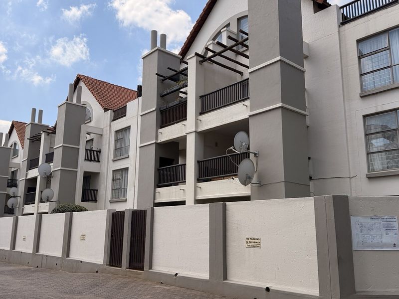Exterior view of a multi-story apartment complex featuring balconies, tiled roofing, and a secure perimeter wall with a paved driveway.