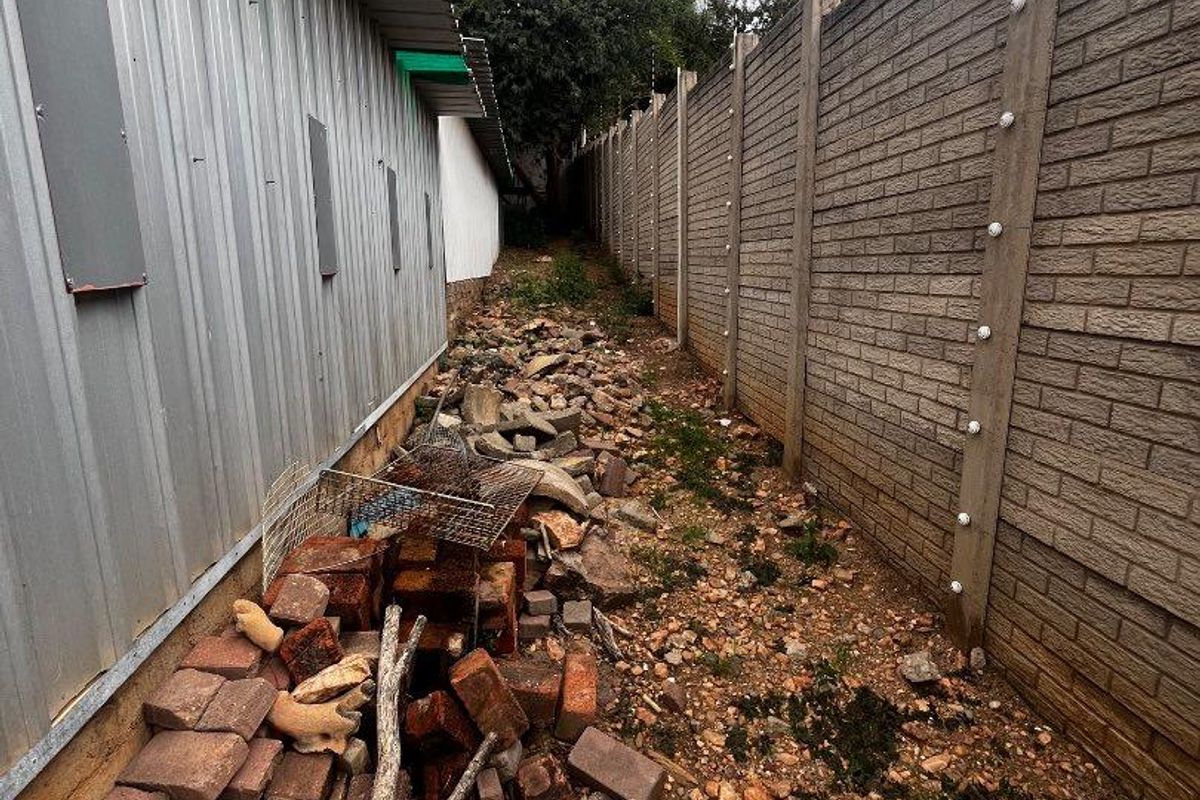 A narrow side passage between the building and the boundary wall, featuring corrugated metal cladding and an electric fence.