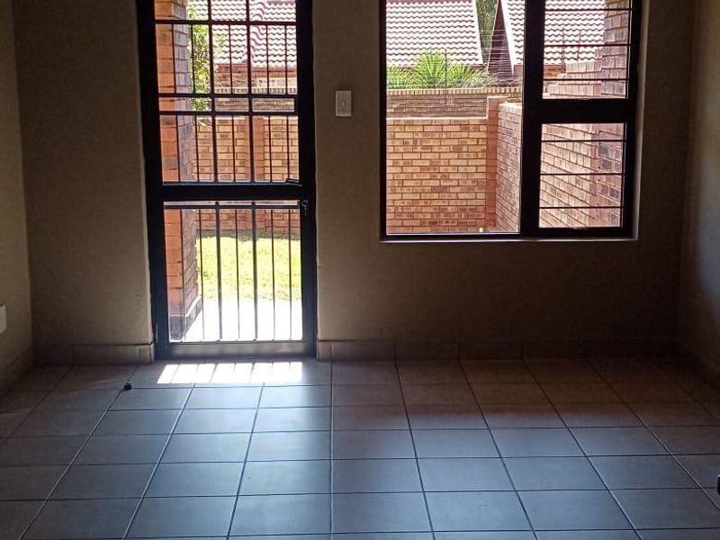 An empty living room featuring tiled flooring, a security gate, and large windows with burglar bars, providing access to the rear exterior area.