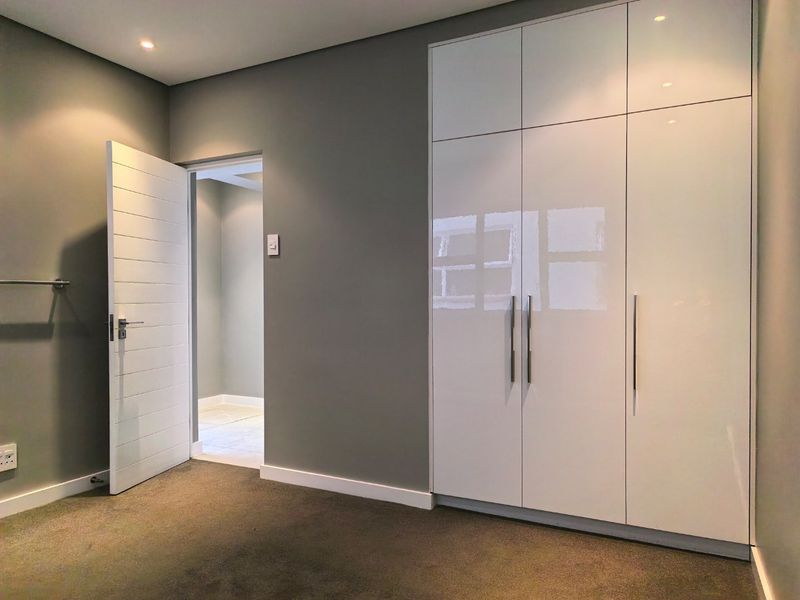 A modern, empty bedroom featuring sleek white built-in cupboards, neutral grey walls, plush carpeting, and recessed downlighting.