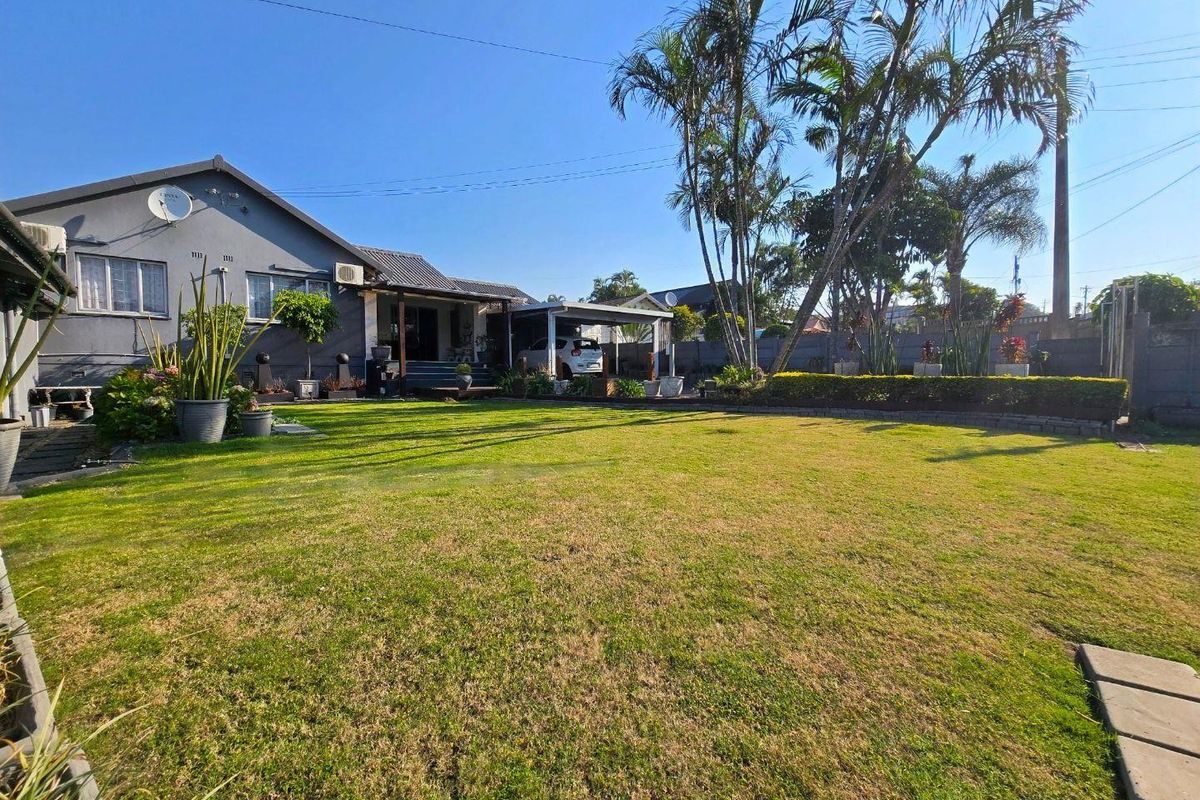 A spacious rear garden featuring a well-maintained lawn, mature palm trees, a paved walkway, and an attached carport area.
