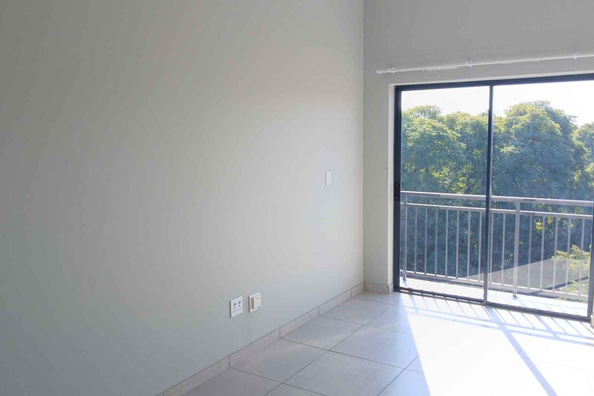 A bright, empty bedroom featuring neutral tiled flooring and a large sliding glass door that opens onto a balcony with leafy views.