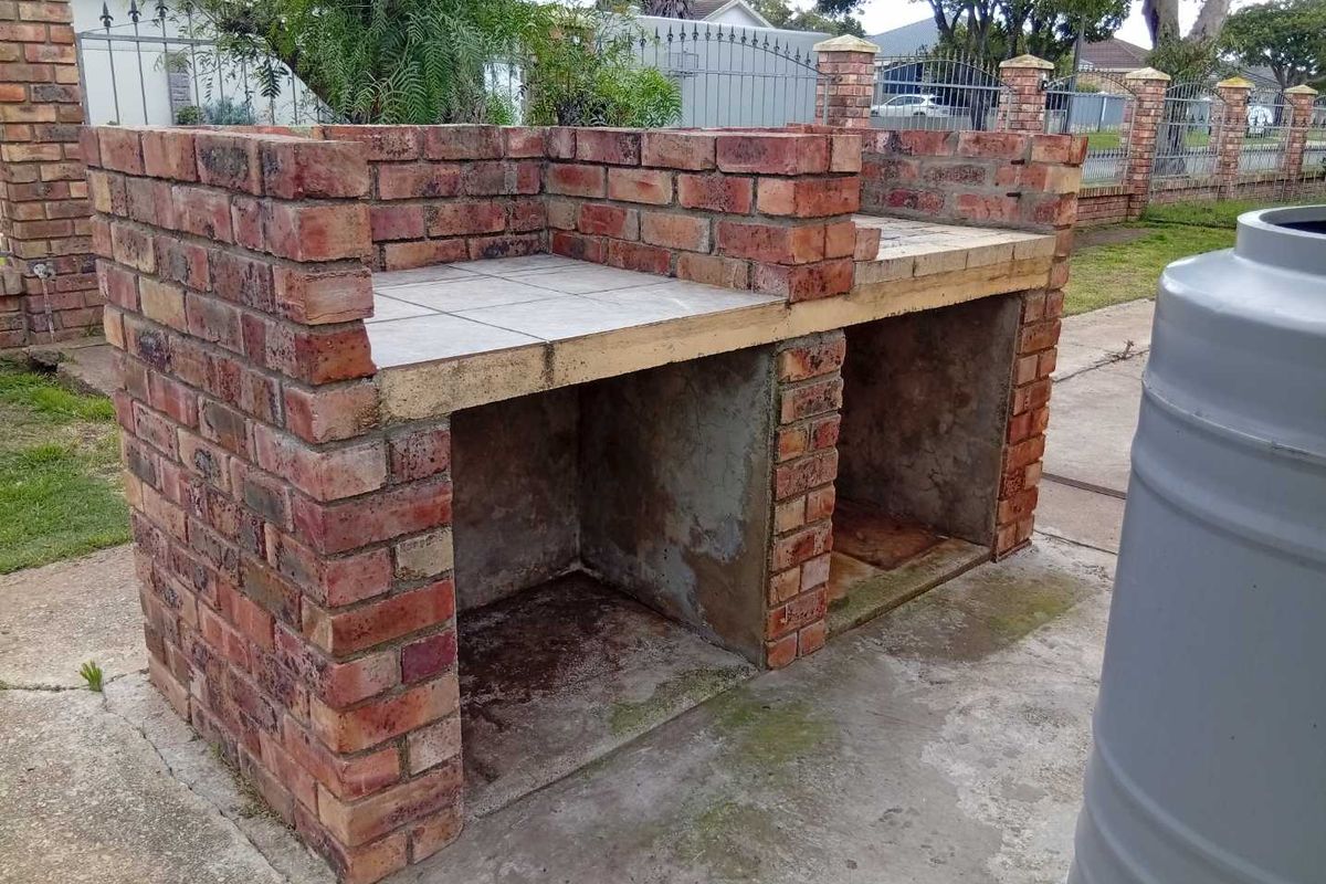 A built-in brick braai area located in the backyard, featuring a tiled prep surface and concrete base, showing signs of weathering.