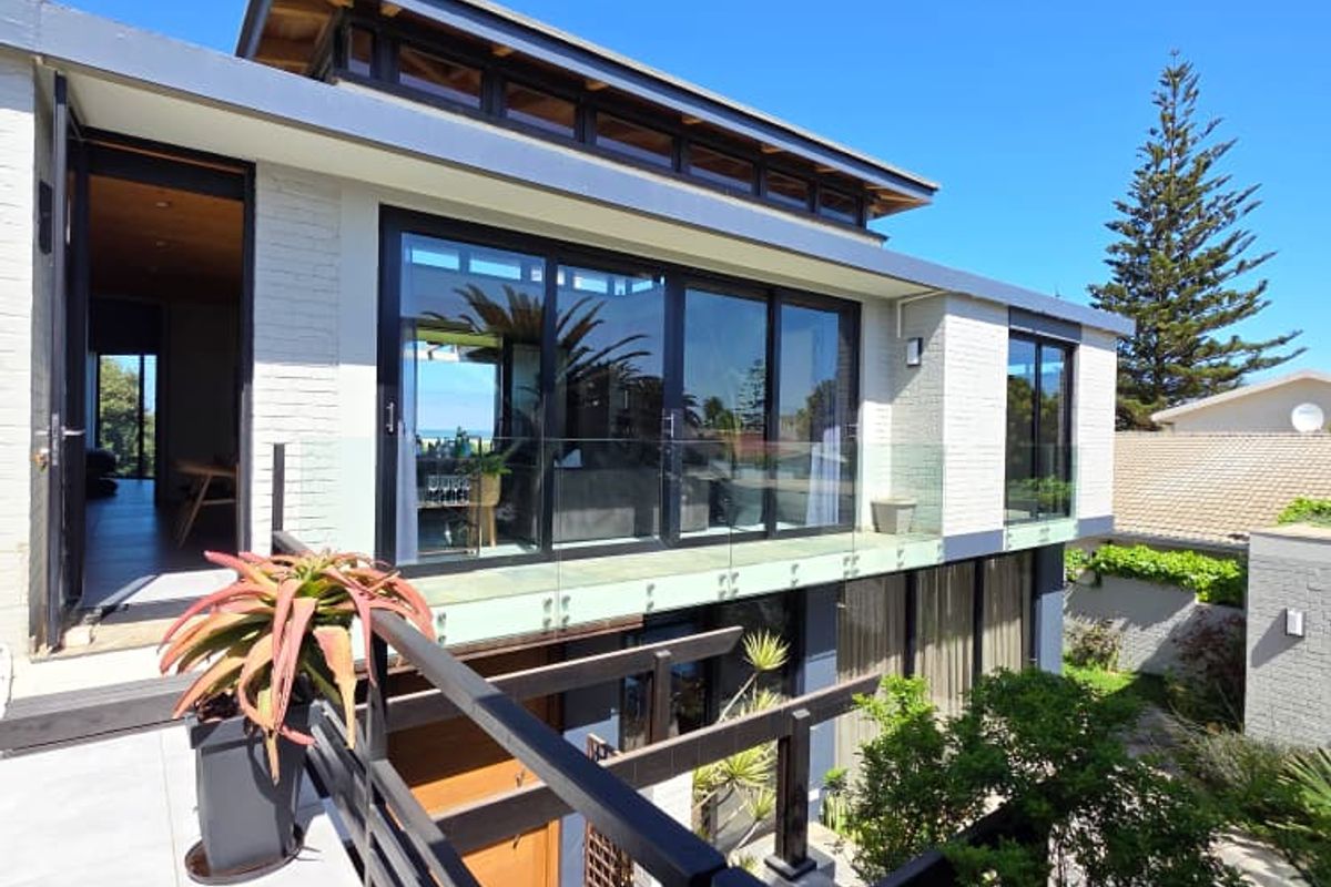 Modern multi-level rear exterior featuring a spacious tiled balcony with glass balustrades, large sliding doors, and a timber pergola overlooking the garden.