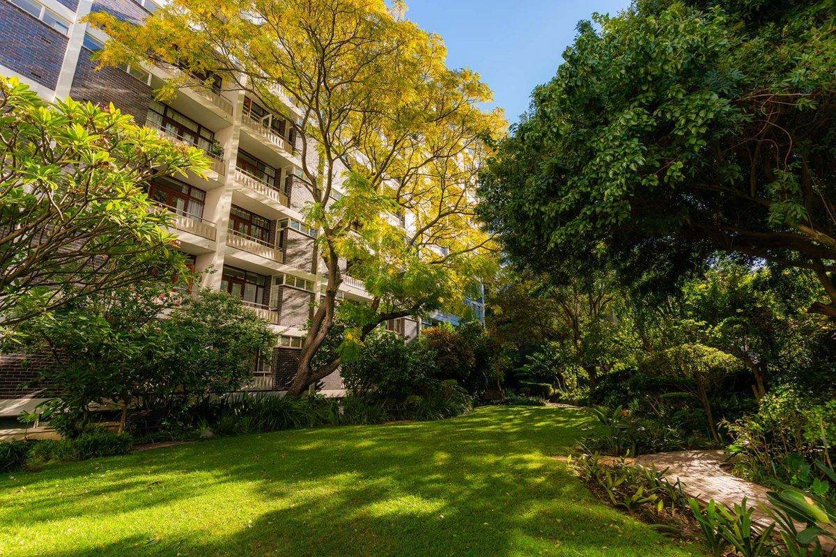 A view of the apartment building exterior featuring a well-maintained garden, mature trees, and private balconies overlooking the green space.