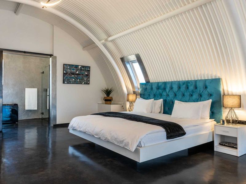 Modern bedroom featuring a unique curved corrugated metal ceiling, polished concrete floors, and a skylight, with an integrated en-suite bathroom.