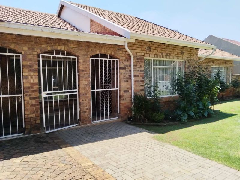 Front elevation of a face brick house featuring a tiled roof, paved driveway, and secure metal entrance gates with established garden beds.