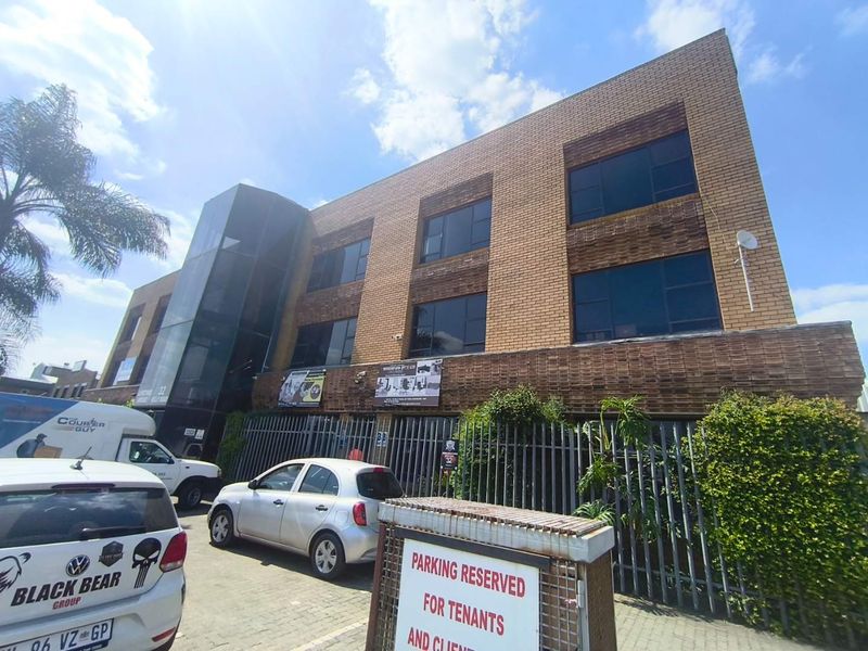 A three-story commercial office building featuring a brick facade, glass stairwell enclosure, and secure perimeter fencing with an on-site paved parking area.