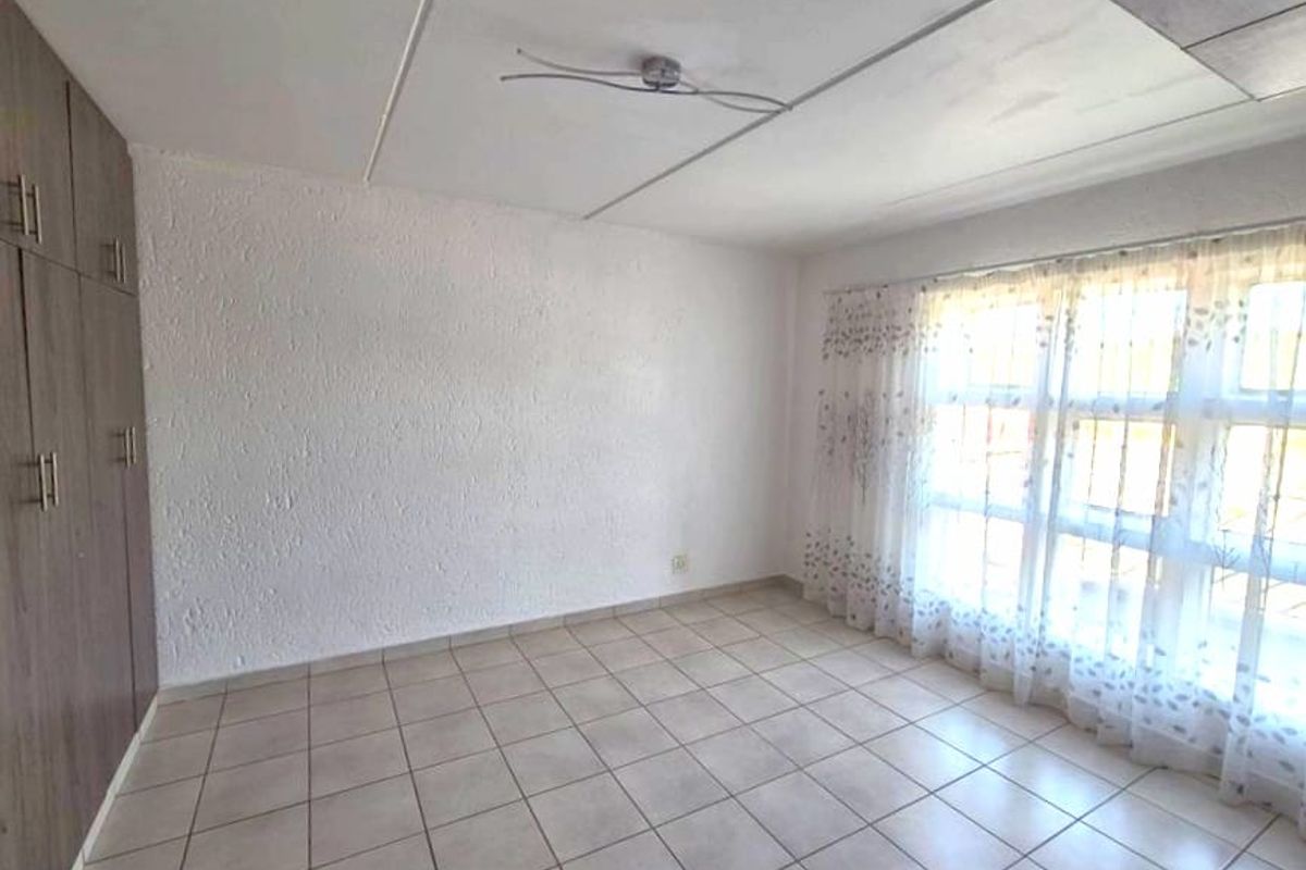 A bright bedroom featuring built-in wooden cupboards, tiled flooring, and large windows with security bars.