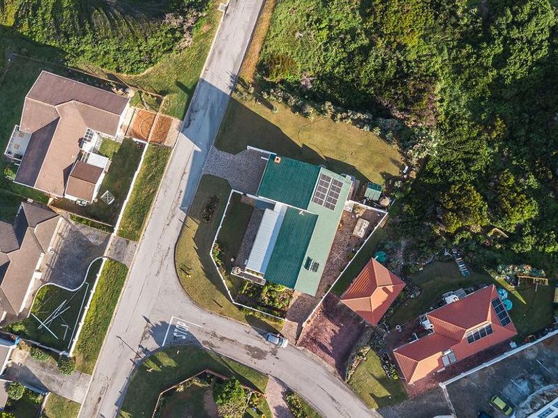 Aerial view of a residential property featuring a green pitched roof with solar panels, a paved driveway, and surrounding landscaped garden areas.